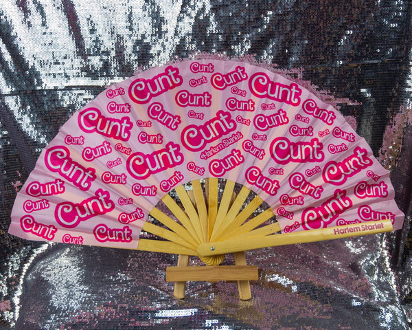 A Bamboo & Silk handheld folding fan that is Pink with a Hot Pink Cunt print pattern is spread open on a silver sequin background.